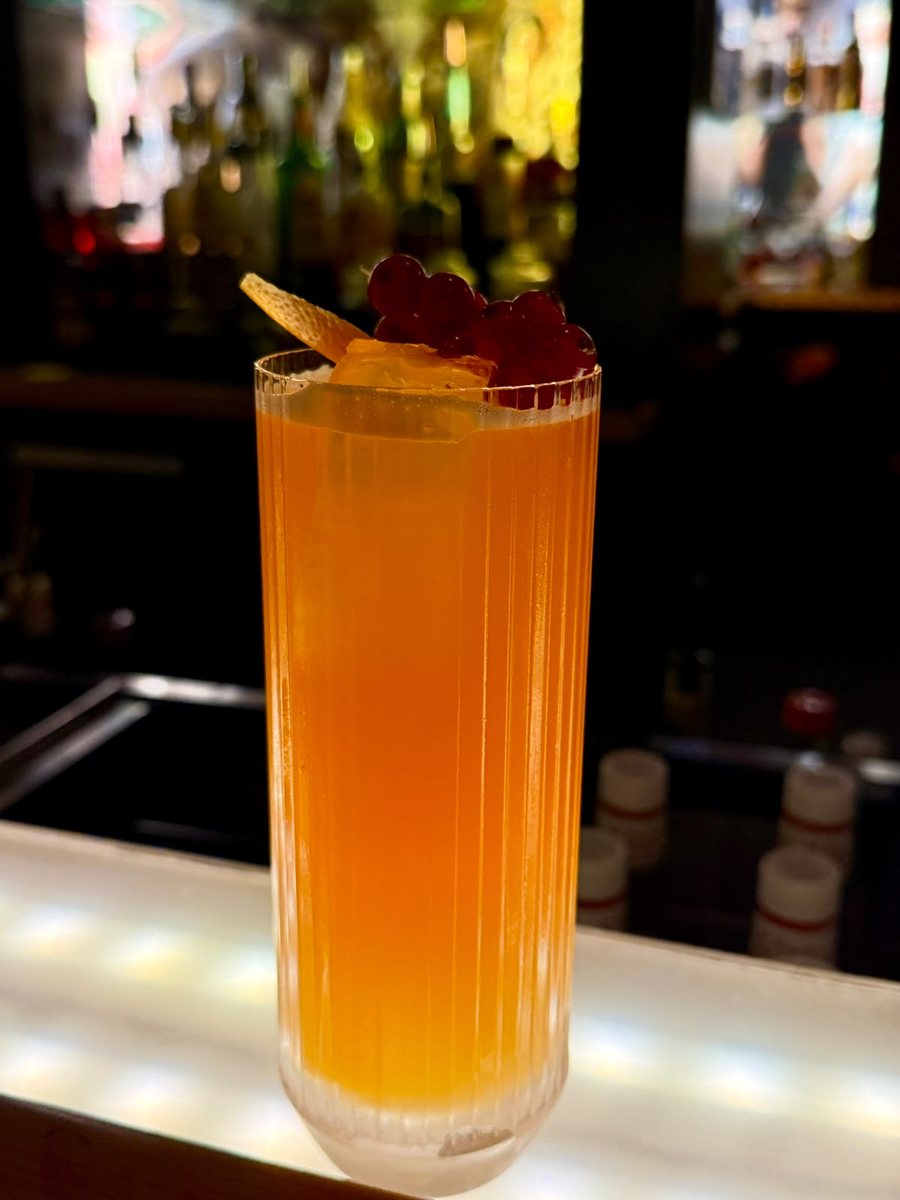 Penumbra im gerippten Longdrinkglas, orange, mit Zitronenzeste und roten Johannisbeeren
