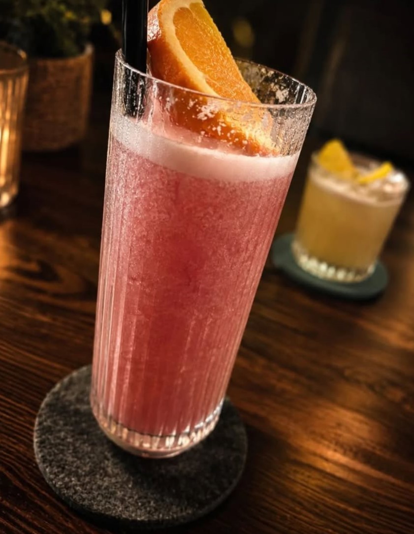 Rosafarbener Longdrink mit Orangenscheibe im Rillenglas auf dem Holztresen