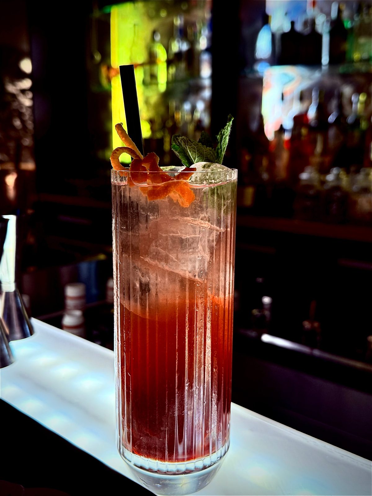Roter Longdrink im gerippten Glas mit Orangenzeste, Minze und schwarzem Strohhalm vor dem beleuchteten Flaschenregal